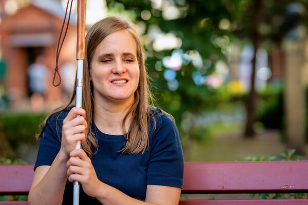 A visually impaired woman holding a white cane and sitting on a bench in the city