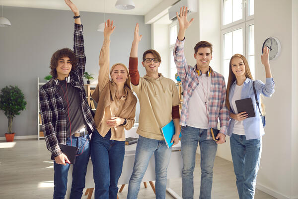 Group of happy smart students standing in classroom and raising hands all together